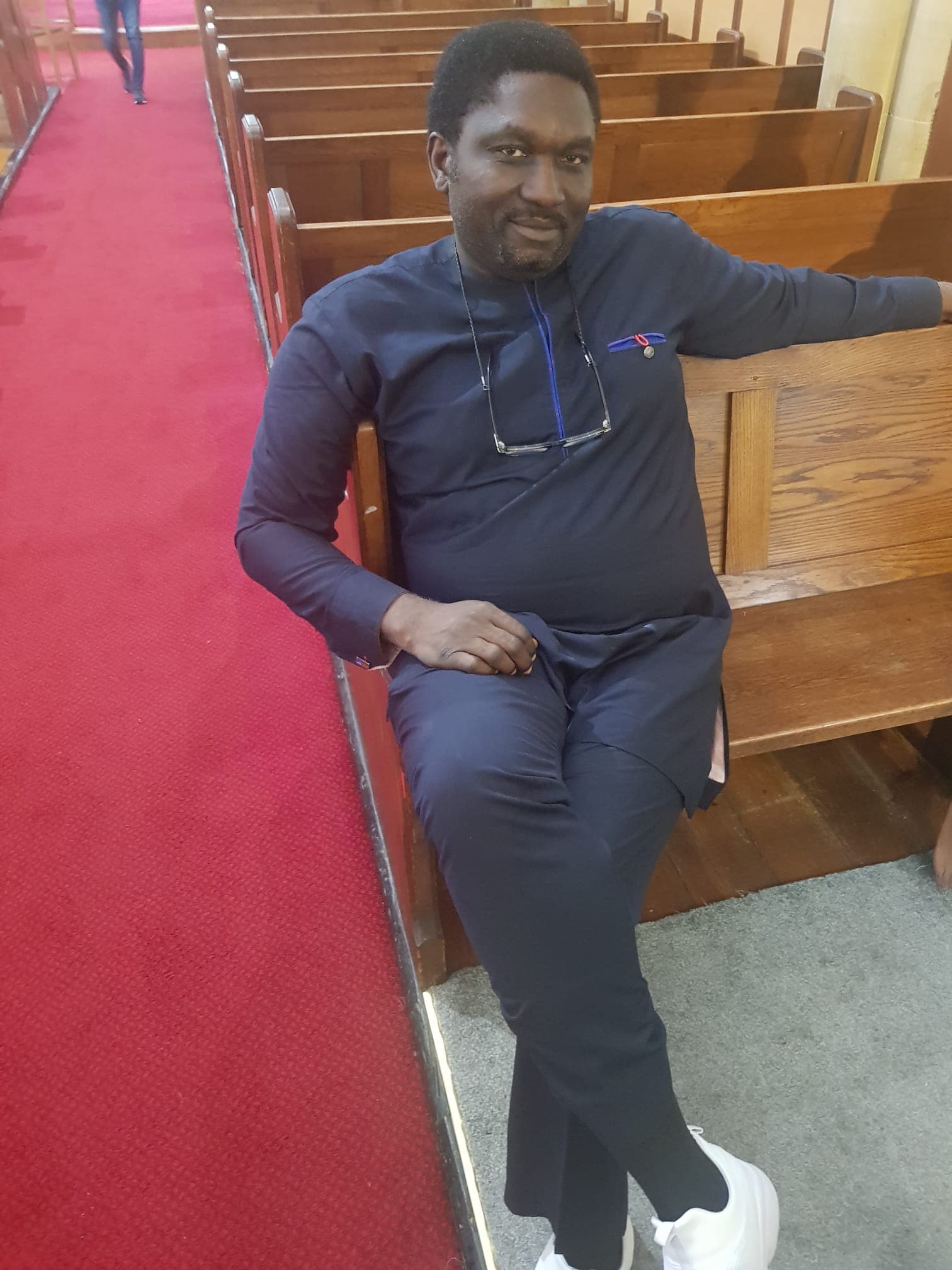 Jide seated in a church pew in a navy kaftan, one arm resting along the back of the bench and his glasses hanging at the collar, half-smiling at the camera.